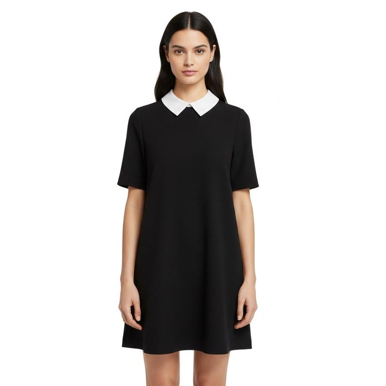 Puede incluir: Un vestido negro con cuello blanco y mangas cortas. El vestido tiene una silueta sencilla en forma de A. La modelo lleva el vestido y est&aacute; de pie frente a un fondo blanco.