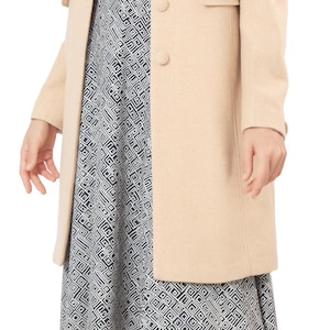 Op de afbeelding: Een beige jas met knopen en kraag, gedragen over een zwart-wit patroon jurk. De outfit wordt gecompleteerd met zwarte sandaaltjes met bandjes. De jas is een neutrale kleur.