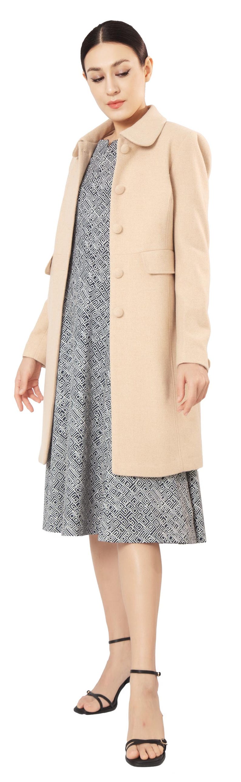 Op de afbeelding: Een beige jas met knopen en kraag, gedragen over een zwart-wit patroon jurk. De outfit wordt gecompleteerd met zwarte sandaaltjes met bandjes. De jas is een neutrale kleur.