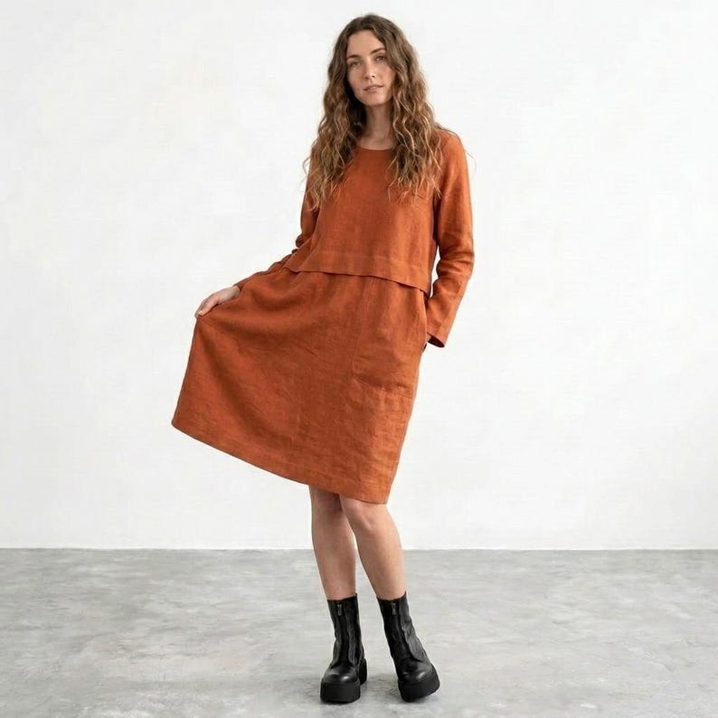 May include: A long-sleeved, knee-length dress in a solid burnt orange color. The garment features a round neckline, a layered bodice design, and a relaxed, A-line skirt silhouette. The fabric has a visible textured weave, and the dress is paired with black platform ankle boots.