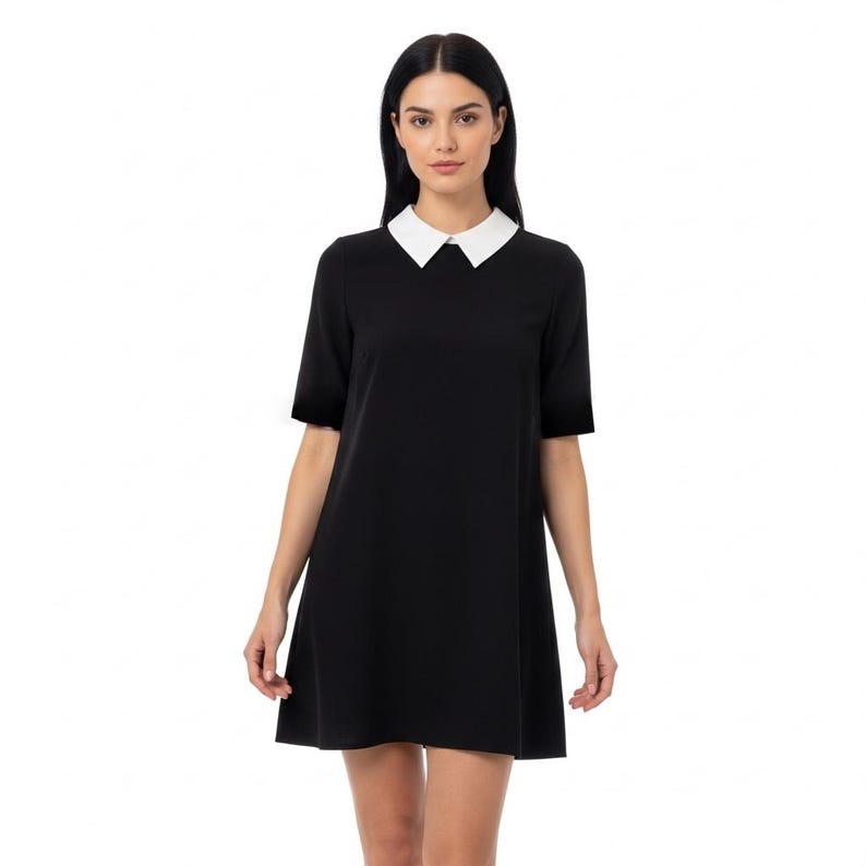Puede incluir: Vestido negro con cuello blanco. El vestido tiene mangas cortas y una silueta sencilla en forma de A. La modelo lleva el vestido, que llega por encima de las rodillas.