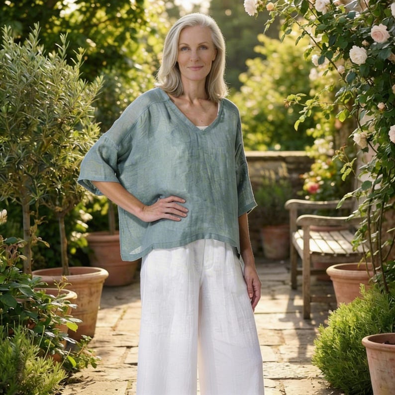 May include: A woman wearing a light green linen top and white wide-leg pants. The top has a V-neck and short sleeves. The pants are high-waisted. The woman is standing in a garden setting with plants and flowers.
