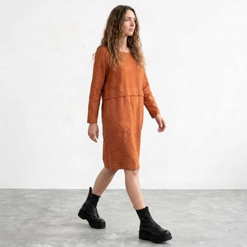 May include: A knee-length, long-sleeved dress in a solid burnt orange color. The garment features a round neckline and a horizontal seam across the chest, creating a layered appearance. The dress is paired with black platform ankle boots.
