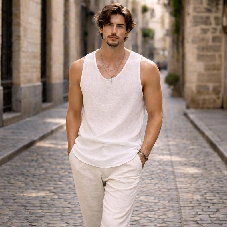 May include: A man wearing a white tank top and light beige pants walks down a cobblestone street. The tank top has a scoop neck and the pants have a relaxed fit. He is also wearing a silver necklace and bracelet.