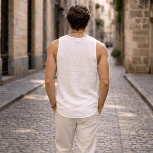 May include: A person wearing a white tank top and beige pants, walking away from the camera on a cobblestone street. The tank top is sleeveless and appears to be made of a lightweight fabric. The pants are a light beige color.