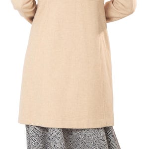 Op de afbeelding: Een beige, knielange jas met een getailleerde pasvorm. De jas wordt gecombineerd met een rok met zwart-witte patronen. Het model draagt zwarte sandalen met hoge hakken. De afbeelding is van achteren genomen.