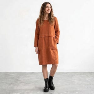 May include: A knee-length, long-sleeved dress in a solid burnt orange color. The garment features a round neckline, a horizontal seam across the chest, and a straight-cut skirt. The fabric has a visible textured weave, and the dress is paired with black boots.