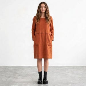 May include: A knee-length, rust-colored dress with long sleeves, a round neckline, and a horizontal seam across the chest. The garment features a straight silhouette with two visible side pockets and is paired with black ankle boots.