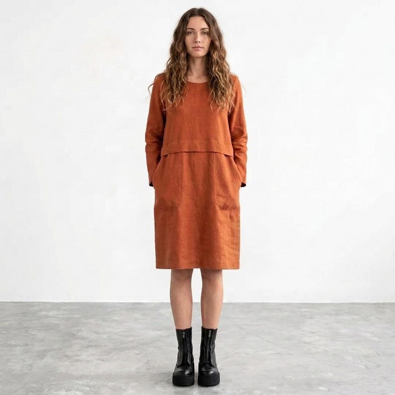 May include: A knee-length, rust-colored dress with long sleeves, a round neckline, and a horizontal seam across the chest. The garment features a straight silhouette with two visible side pockets and is paired with black ankle boots.