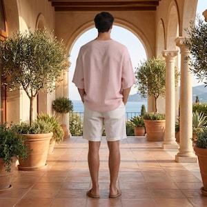 May include: A person wearing a light pink linen shirt and white shorts. The outfit is paired with brown sandals. The person is standing on a tiled patio with potted plants and a view of the ocean.
