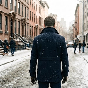 Puede incluir: Un abrigo de lana azul marino con cuello de piel marr&oacute;n, usado por una persona que camina por una calle adoquinada cubierta de nieve. La persona lleva guantes de cuero marr&oacute;n. Los edificios bordean la calle.