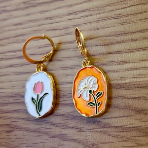 Könnte beinhalten: Ein Paar goldfarbene Ohrringe mit ovalen Anhängern. Ein Anhänger zeigt eine weiße Blume mit einem grünen Stiel auf orangefarbenem Hintergrund. Der andere Anhänger zeigt eine rosa Blume mit einem grünen Stiel auf weißem Hintergrund.