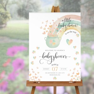 May include: A baby shower sign on a wooden easel. The sign features a watercolor design with a rainbow, pot of gold, and hearts. Text includes "a little lucky charm is on the way" and "baby shower" with the date and time.
