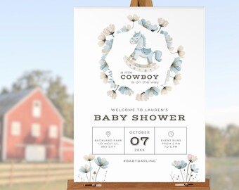 May include: A baby shower welcome sign on a wooden easel. The sign features a blue rocking horse illustration surrounded by a floral wreath. Text includes "a little cowboy is on the way" and event details.