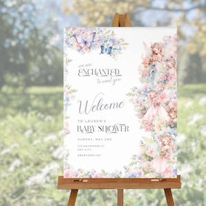Puede incluir: Un cartel de bienvenida blanco con un borde floral y dos hadas. El texto dice "We are Enchanted to meet you. Welcome to Lauren's Baby Shower. Sunday, May 2nd. 123 Anywhere St. Any City. #BabyDarling"