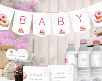May include: Pink and white baby shower decorations with a donut theme. The decorations include a banner that says "BABY", cupcake toppers, water bottle labels, and signs that say "Cupcakes, Do Not Touch These" and "Water, Do Not Touch These".