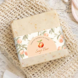 Peut inclure: Un pain de savon rectangulaire avec une étiquette décorative représentant des illustrations de pêches et le texte "FROM MY SHOWER TO YOURS Soap Bar". Le savon est beige clair et semble fait à la main. L'étiquette a un demi-cercle couleur pêche.