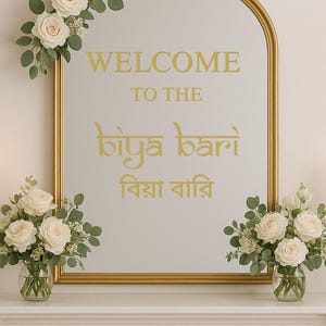 Pode incluir: Espelho com moldura dourada com as palavras "WELCOME TO THE biya bari" em letras douradas. O espelho é decorado com rosas brancas e vegetação. Dois vasos de rosas brancas estão colocados abaixo.