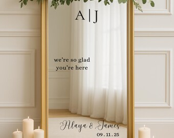 May include: A gold-framed mirror with floral arrangements and candles. The mirror displays the text "A|J", "we're so glad you're here", "Alaya & James", and the date "09.11.25". White roses and greenery adorn the top of the mirror.