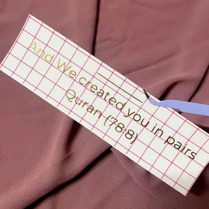 Op de afbeelding: Een wit vel papier met een rasterpatroon en gouden letters met de tekst "And We created you in pairs Quran (78:8)"