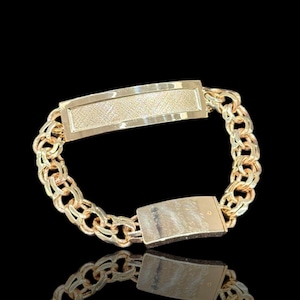 May include: A gold-colored bracelet with a textured rectangular plate and a double-link chain design. The bracelet is displayed against a black background, highlighting its reflective surface and intricate details. The plate is approximately 1 inch long.