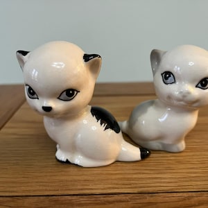 May include: Two ceramic cat figurines, one white with black markings and the other gray with black eyes. Both have a cartoonish style.