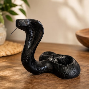 May include: A black cobra figurine with a textured surface, a raised hood, and a thin, protruding tongue. The snake is depicted in a coiled position with its head upright, featuring small, light-coloured eyes and a dark, matte finish.