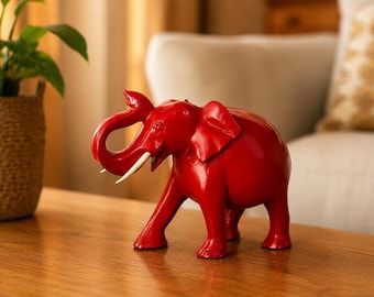 Könnte beinhalten: Eine leuchtend rote Elefantenfigur, die in einer nach vorne gerichteten Position steht, wobei der Rüssel nach oben gebogen ist. Der Elefant hat eine glatte, glänzende Haut und zwei markante, geschwungene weiße Stoßzähne, die aus dem Maul ragen.
