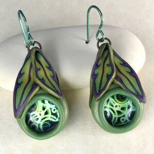 May include: A pair of green and purple dangle earrings with a green and white patterned circle in the center. The earrings are made of polymer clay and have a wire hook.
