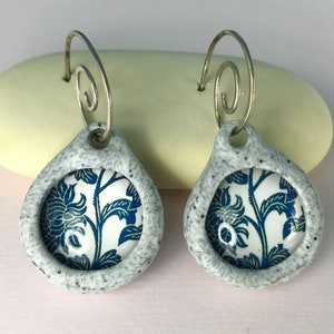 May include: A pair of dangle earrings with a white ceramic base and a blue and white floral design. The earrings have a silver wire hook.