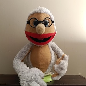 May include: A plush puppet featuring a tan face, a bright red mouth, and white fuzzy hair. The puppet wears round black-rimmed glasses and has white fuzzy arms and hands. The puppet is constructed with a tan torso and is mounted on a green base.