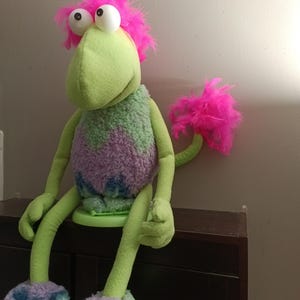 May include: A plush creature puppet with bright pink hair and a matching pink feather tuft on its tail. The body is light green with a textured purple and blue midsection. It has large white eyes, long green arms and legs, and purple feet.