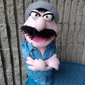 May include: A handmade puppet with a pink face, large eyes, and a black mustache. It wears a gray hat and a blue Hawaiian shirt. The puppet's arms are crossed, and it is set on a blue table against a textured wall.