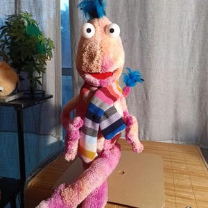 Peut inclure: Une marionnette en peluche avec un corps rose et pêche, de grands yeux globuleux et une bouche rouge. Elle porte une écharpe rayée colorée et a des accents de plumes bleues. La marionnette est assise sur une surface en bois.