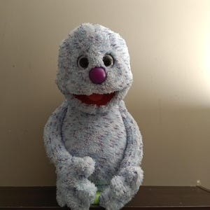 May include: A plush puppet featuring light blue fur with dark blue speckles, large round black eyes, a prominent purple nose, and an open mouth with a red interior. The puppet has long arms and legs, and is constructed from soft, textured material.