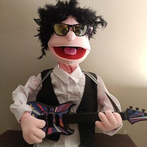 May include: A puppet with black hair, sunglasses, and a pink face, wearing a white shirt, black vest, and holding a colorful toy guitar. The guitar has a black neck and a body with a star and stripe design.