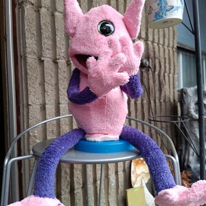 May include: A pink plush creature with large ears, green eyes, and outstretched hands. It has purple arms and legs, and is sitting on a metal chair. The creature's mouth is open, and it appears to be a puppet or toy.