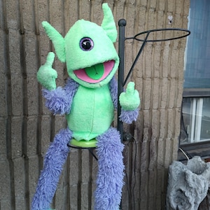 May include: A plush green and purple creature with large ears, a wide mouth, and a tongue. The creature is seated on a black metal stand, with its arms raised, displaying its fingers. The creature's body is green, with purple arms and legs.