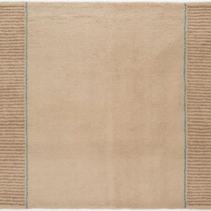 May include: A rectangular beige rug with a soft, textured surface. The rug features a striped border in a darker tan color on both sides, with a thin blue line separating the border from the main body of the rug. The edges have small tassels.