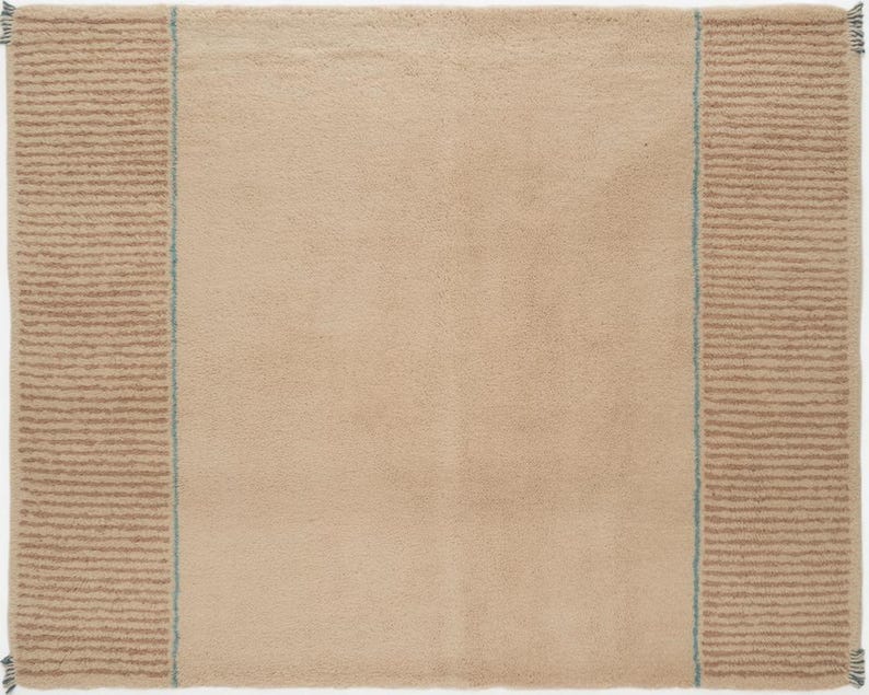 May include: A rectangular beige rug with a soft, textured surface. The rug features a striped border in a darker tan color on both sides, with a thin blue line separating the border from the main body of the rug. The edges have small tassels.