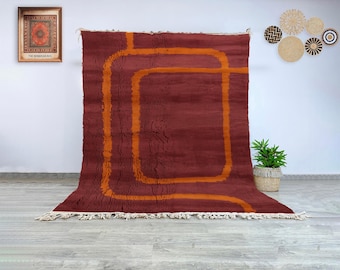 Könnte beinhalten: Ein großer, rechteckiger, kastanienbrauner Teppich mit einem geometrischen, orangefarbenen Design. Der Teppich hat einen Fransenbesatz am unteren Rand und wird vor einer weißen Wand präsentiert. Der Teppich ist ein dekoratives Wohnaccessoire.
