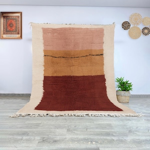 May include: A rectangular rug with a cream-colored border and a central design of horizontal bands in shades of pink, tan, and burgundy. The rug has a fringed edge and a textured appearance, suitable for home decor.