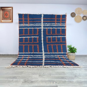 Puede incluir: Una alfombra azul rectangular grande con un patrón geométrico de líneas y formas naranjas y blancas. La alfombra tiene flecos a lo largo del borde inferior. La alfombra se exhibe contra una pared blanca.
