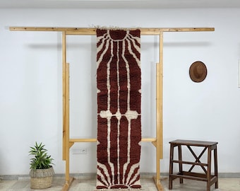 May include: A maroon and cream-coloured rectangular rug with a geometric design hangs on a wooden rack. The rug has a sunburst pattern at the top and bottom, with vertical stripes in between. A small wooden stool and a potted plant are also visible.