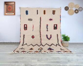 May include: A large, cream-colored rug with a geometric pattern. The rug features colorful, abstract shapes in red, blue, green, and brown. The rug has a fringed edge and is displayed against a white wall.