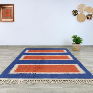 Puede incluir: Una alfombra rectangular azul con tres dise&ntilde;os rectangulares naranjas. La alfombra tiene un borde y flecos color crema. La alfombra est&aacute; sobre un suelo de madera.