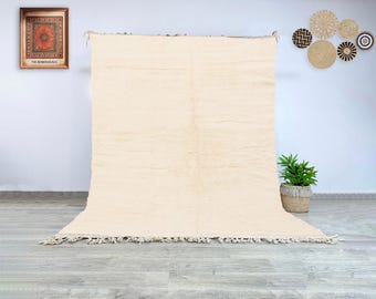 May include: A large, cream-colored rug with a textured surface and fringed edges. The rug is displayed against a white wall, with a framed artwork and decorative wall hangings in the background. The rug is a neutral color and would complement many interior design styles.