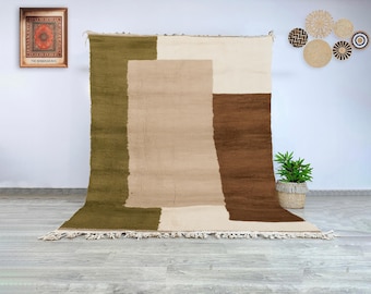 May include: A large, rectangular rug with a geometric design. The rug features blocks of olive green, beige, cream, and brown. The rug has a fringed edge and is displayed against a white wall.