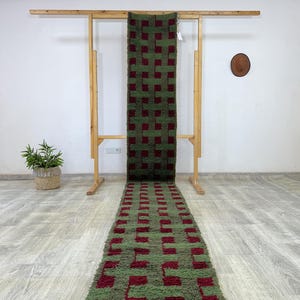 May include: A long, rectangular rug with a green and burgundy checkered pattern. The rug is displayed on a wooden rack, showcasing its design. The rug's texture appears soft and plush, suitable for a hallway or entryway.