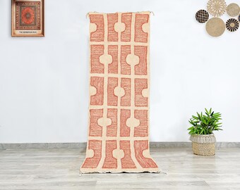 May include: A long, rectangular rug with a geometric pattern in beige and terracotta. The rug is displayed vertically against a white wall, showcasing its design and texture. The rug's design features a repeating pattern of rectangles and circles.